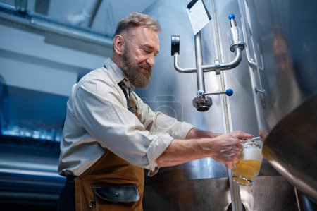 Téléchargez les photos : Brewery master quality tasting of craft beer at production facility. Man brewer pouring freshly brewed ale or lager into glass from the metal tank - en image libre de droit