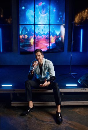 Téléchargez les photos : Male singer performing at music concert. Handsome emotional musician singing song in microphone sitting on stage of nightclub - en image libre de droit