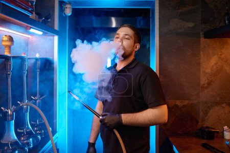 Photo for Young man smoking at hookah bar while preparing equipment for client. Guy emitting puffs of smoke from opened mouth - Royalty Free Image