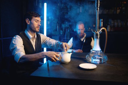 Foto de Buenos amigos reunidos en el salón de narguile. Dos tipos fumando shisha y bebiendo té en la cafetería o el bar. Concepto de relajación - Imagen libre de derechos