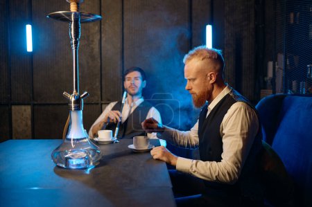 Foto de Buenos amigos reunidos en el salón de narguile. Dos tipos fumando shisha y bebiendo té en la cafetería o el bar. Concepto de relajación - Imagen libre de derechos