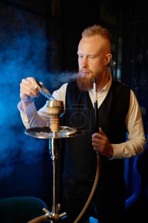 Photo for Stylish bearded man mixing tobacco in hookah while smoking. Smoker vaping shisha pipe during recreation time at night bar - Royalty Free Image