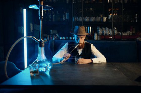 Photo for Portrait of stylish bearded man with hat smoking hookah. Fashion guy sitting at table and blowing steam vortexes - Royalty Free Image