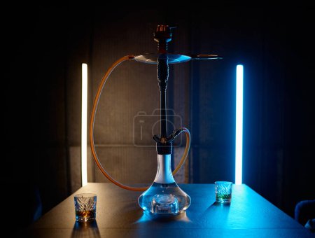 Foto de Hookah with glass flask and metal bowl shisha on table with two portion of brandy in dark room - Imagen libre de derechos