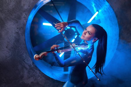 Photo for Young artistic woman performer playing violin in atmospheric studio. Neon light, futuristic industrial design - Royalty Free Image