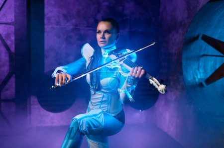 Téléchargez les photos : Jeune femme jouant du violon solo sur scène éclairée par une lumière au néon dynamique sur fond industriel - en image libre de droit