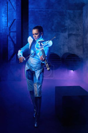 Téléchargez les photos : Young woman playing violin solo on stage illuminated with dynamic colored neon light over industrial background - en image libre de droit