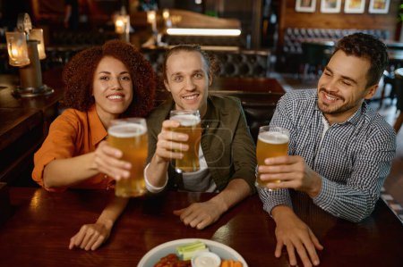 Photo for Cheerful friends drinking draft beer at bar table in sports pub. Group of young people with raising beer glasses looking at camera - Royalty Free Image
