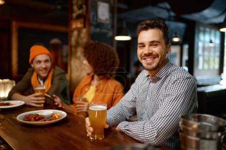 Foto de Portrait of smiling sports fan sitting counter desk. Handsome cheerful man drinking craft beer and looking at camera while rest with friends in sport bar - Imagen libre de derechos