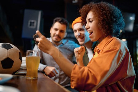 Foto de Cheerful friends having fun watching football game on smartphone at sport bar. Extremely emotional young people drinking draft beer at bar counter in pub - Imagen libre de derechos