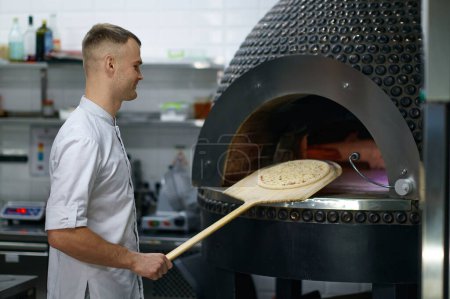 Photo for Professional baker chef making traditional italian pizza in oven. Man in uniform holding long shovel over modern restaurant kitchen interior - Royalty Free Image