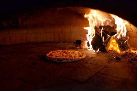 Foto de Pizza is cooked in traditional wood oven with open fire. Italian fast food preparation in professional stove with burning firewood - Imagen libre de derechos