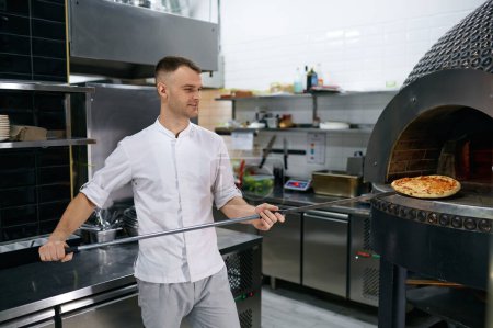 Photo for Young male chef holding freshly baked pizza on long shovel. Selective focus of hot Italian fast food - Royalty Free Image