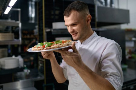 Photo for Satisfied male chef with smile on face enjoying smell of just baked pizza. Delicious handmade Italian pastry preparation - Royalty Free Image