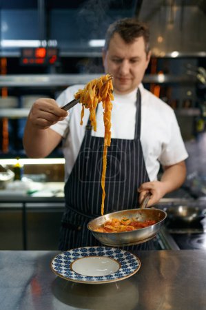Photo for Professional chef serving freshly cooked pasta on plate. Portrait of mature cooker dressed in apron holding spaghetti on fork - Royalty Free Image