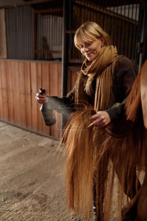 Foto de Horse owner spraying stallion with conditioner during grooming and cleaning before riding. Taking care of domestic animal in livestock - Imagen libre de derechos