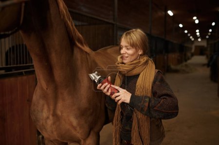Foto de Young satisfied woman grooming horse using electric shaver in ranch stable. Concept of veterinary, equine breeding and taking care of the horse - Imagen libre de derechos