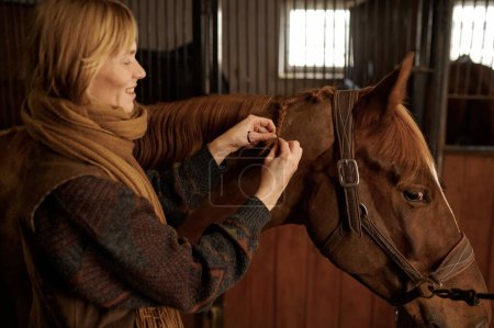 Foto de Portrait of woman braiding horse mane while standing in stable of riding club. Animal grooming and hairstyling concept - Imagen libre de derechos