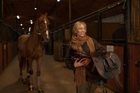 Foto de Female horse rider walking with harness in stable. Young adult woman rancher going with saddle in hands and smiling - Imagen libre de derechos
