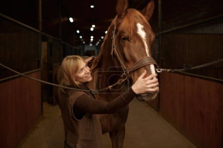 Foto de Horsewoman petting horse stroking animal muzzle over stable interior. Friendship, loving and taking care of animal - Imagen libre de derechos