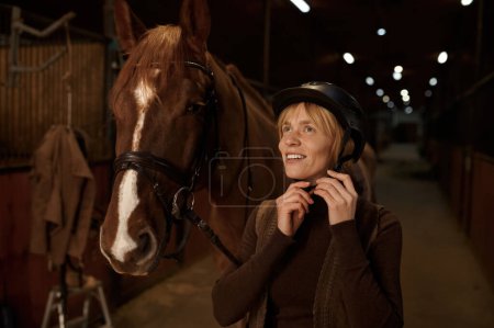 Foto de Mujer jinete poniendo casco en la cabeza de pie delante del caballo arnés en el establo del club de equitación moderno - Imagen libre de derechos