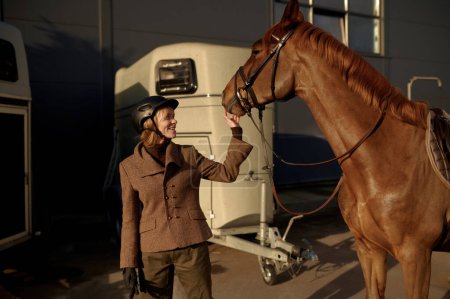 Foto de Joven jinete adulto sonriente mujer acariciando arnés nariz de caballo mostrando amor y cuidado. Concepto equino de amistad - Imagen libre de derechos