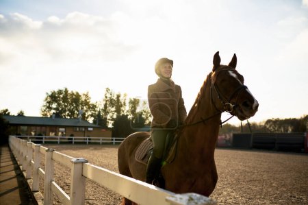 Foto de Attractive smiling female horse rider training in outdoor paddock. Riding club and equine caring concept - Imagen libre de derechos