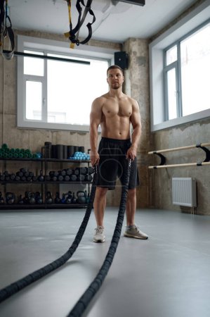 Foto de Young strong man pulling training sports rope front view. Male athlete doing workout exercises at gym - Imagen libre de derechos