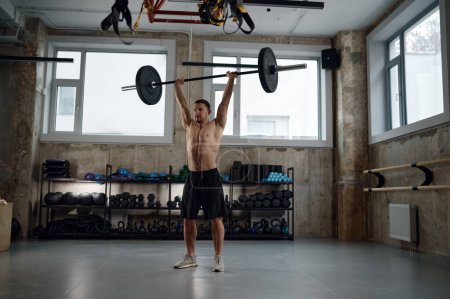 Photo for Muscular guy lifting barbell above head standing at gym. Young athlete under physical exertion using sports weight equipment during fitness workout - Royalty Free Image