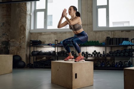 Photo for Strong muscular woman practicing sports jumping on wooden fitness box at gym. Healthy female trainer in sportswear training alone. - Royalty Free Image