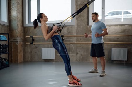 Foto de Woman training exercising her muscles with help of suspension straps under professional sport coach control in gym - Imagen libre de derechos