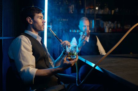 Foto de Dos hombres jóvenes y elegantes relajarse y fumar narguile en el bar. Amigos de negocios fuman de pipa de shisha y beben en restaurante - Imagen libre de derechos