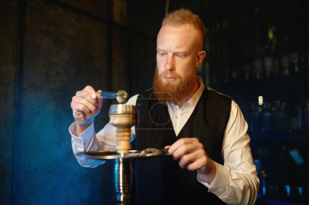 Photo for Stylish bearded man mixing tobacco in hookah while smoking. Smoker vaping shisha pipe during recreation time at night bar. Closeup view - Royalty Free Image