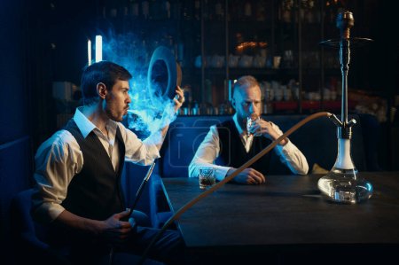 Téléchargez les photos : Retro well-dressed gangsters smoking hookah in bar. Serious handsome guys having business meeting while spending time at nightclub - en image libre de droit