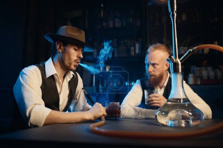Photo for Retro dressed gangsters smoking hookah in bar. Serious handsome guys having business meeting while spending time at nightclub - Royalty Free Image