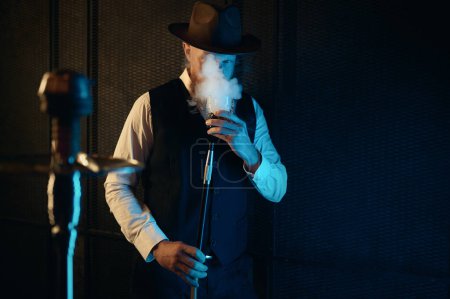 Foto de Vista frontal del hombre en reposo fumando narguile en el lugar de trabajo en casa. Retro chico en sombrero beber relajarse en habitación oscura con iluminación de neón - Imagen libre de derechos