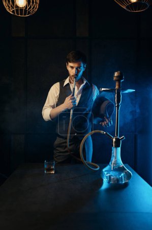 Photo for Retro dressed guy holding hat smoking hookah while standing in clouds of smoke and looking at camera - Royalty Free Image