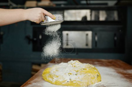Foto de Primer plano panadero manos espolvorear harina sobre la masa cruda. Proceso de preparación de pan en la cocina profesional moderna - Imagen libre de derechos