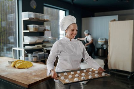 Photo for Portrait of woman baker in uniform carrying tray with cookies shaped from dough and prepared for baking. Pastry production - Royalty Free Image