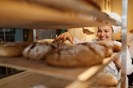 Foto de Mujer feliz panadero en uniforme de control de pan recién horneado en bandeja en la cocina de panadería moderna - Imagen libre de derechos