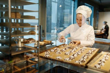 Téléchargez les photos : Boulanger femelle portant un uniforme blanc étalant des biscuits fraîchement cuits. Plaque de cuisson avec pâtisserie sucrée - en image libre de droit