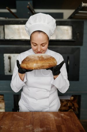 Foto de Joven panadero hembra que huele apetitoso pan fresco sobre el fondo de la casa de panadería comercial moderna. Producción de productos horneados - Imagen libre de derechos
