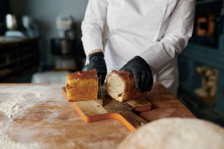 Foto de Joven mujer sonriente panadero chef sosteniendo cuchillo y cortando pan fresco en la mesa de madera. Alimentación saludable y concepto de panadería tradicional - Imagen libre de derechos