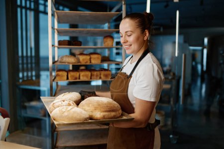 Téléchargez les photos : Jolie femme boulanger tenant variété de pain cuit sur plateau debout sur fond de maison de boulangerie - en image libre de droit