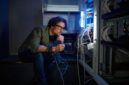 Foto de Ingeniero de red masculino conectando cables en la sala de servidores durante los trabajos de mantenimiento en el centro de datos. Hombre joven eligiendo el hub óptico de fibra óptica para conectar con el equipo - Imagen libre de derechos