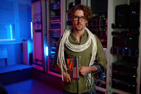 Foto de Retrato de signalman con muchos cables en los hombros y portátil en la mano de pie sobre la moderna sala de servidores en el fondo del centro de datos - Imagen libre de derechos