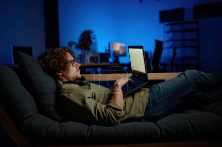 Foto de Hombre administrador de TI descanso en el sofá con ordenador portátil que se encuentra en el sofá en la moderna sala de servidores. Servicio nocturno en concepto de centro de datos - Imagen libre de derechos