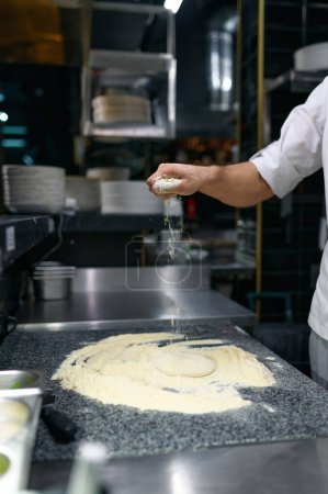 Téléchargez les photos : Gros plan pâtissier chef mains saupoudrer de pâte à farine pour le pain, pâtes ou pizza à la cuisine professionnelle dans le restaurant - en image libre de droit