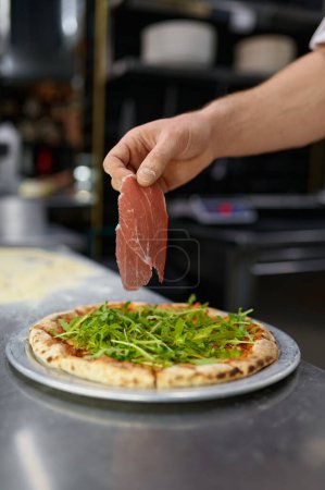 Photo for Delicious pizza preparation process. Closeup chef hand holding slice of bacon over Italian pastry - Royalty Free Image