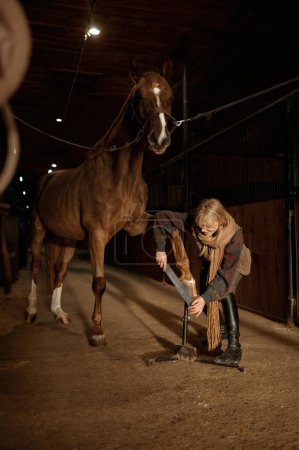 Foto de Young woman making horses hoof manicure, filing and shaping horseshoe. Stallion grooming and cleaning before riding - Imagen libre de derechos